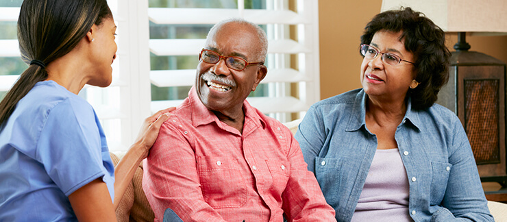 Female care aide places hand on arm of a smiling senior man sitting on the couch with his wife.