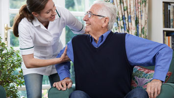 Female care worker helps senior man with paranoia get up from sitting on couch.