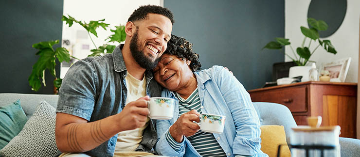 Young man is having coffee with his elderly mom on the sofa at home.