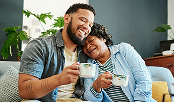 Young man is having coffee with his elderly mom on the sofa at home.